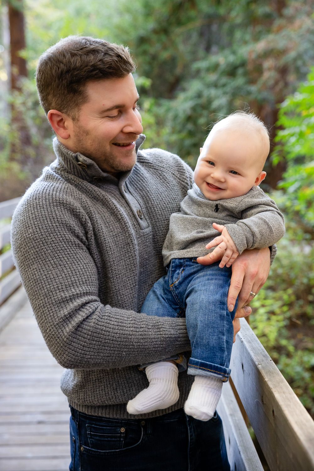 A parent and baby share a joyful moment together while standing on a wooden boardwalk surrounded by forest in Los Altos