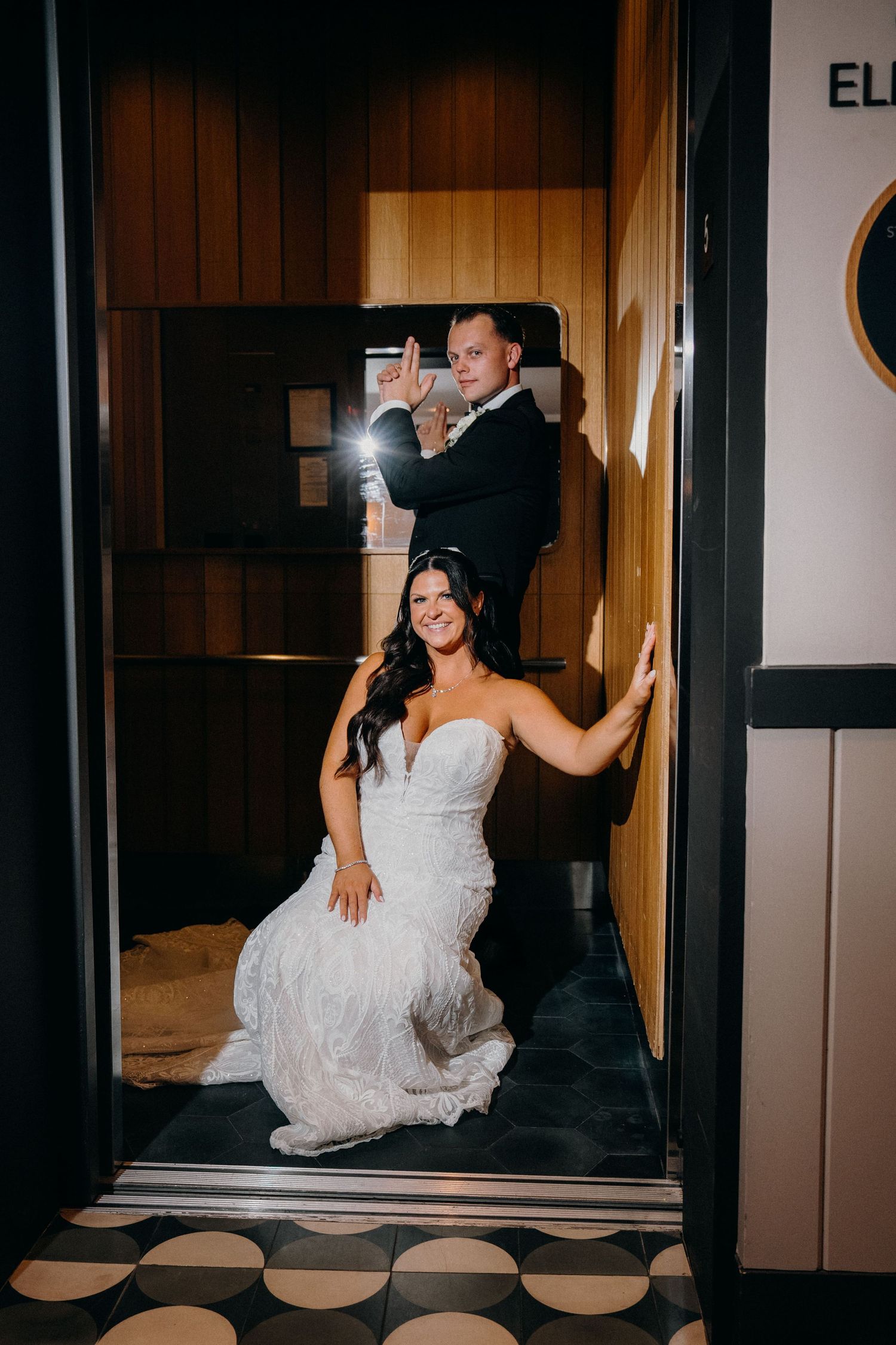 Bride in white wedding dress sits playfully in elevator during wedding photo session.