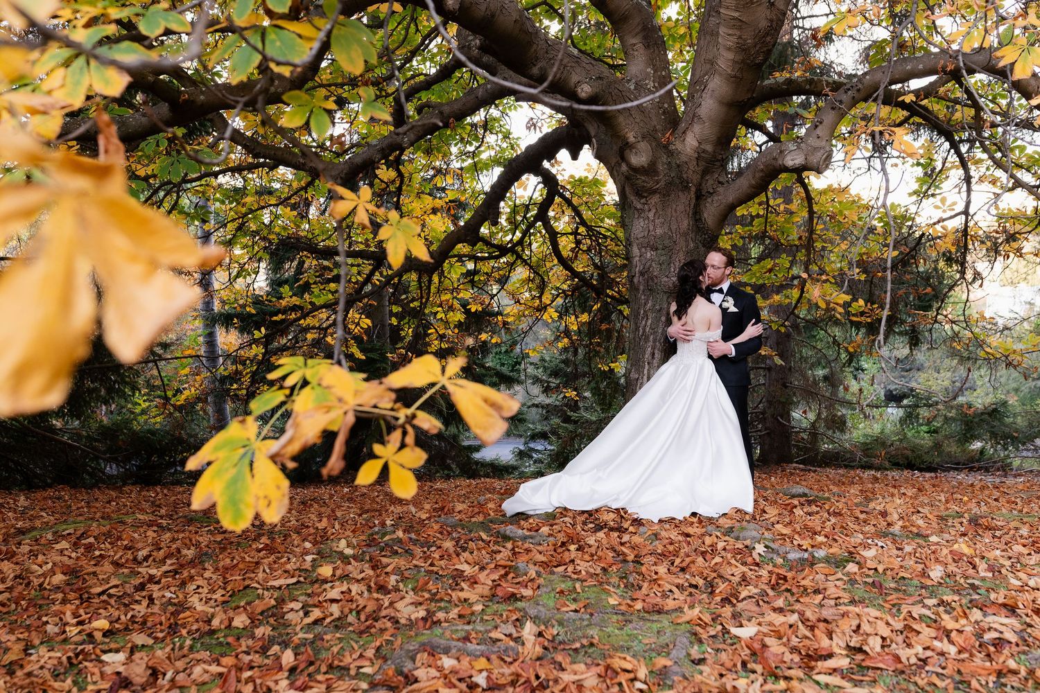 A couple in wedding attire poses for romantic autumn photos among fallen leaves and trees in a park setting.