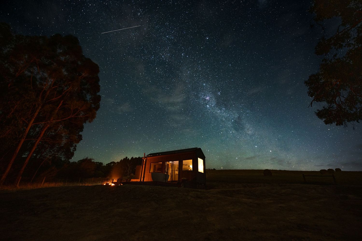 Small cabin under starry night sky with visible Milky Way, campfire glowing outside, shooting star visible above remote Australia