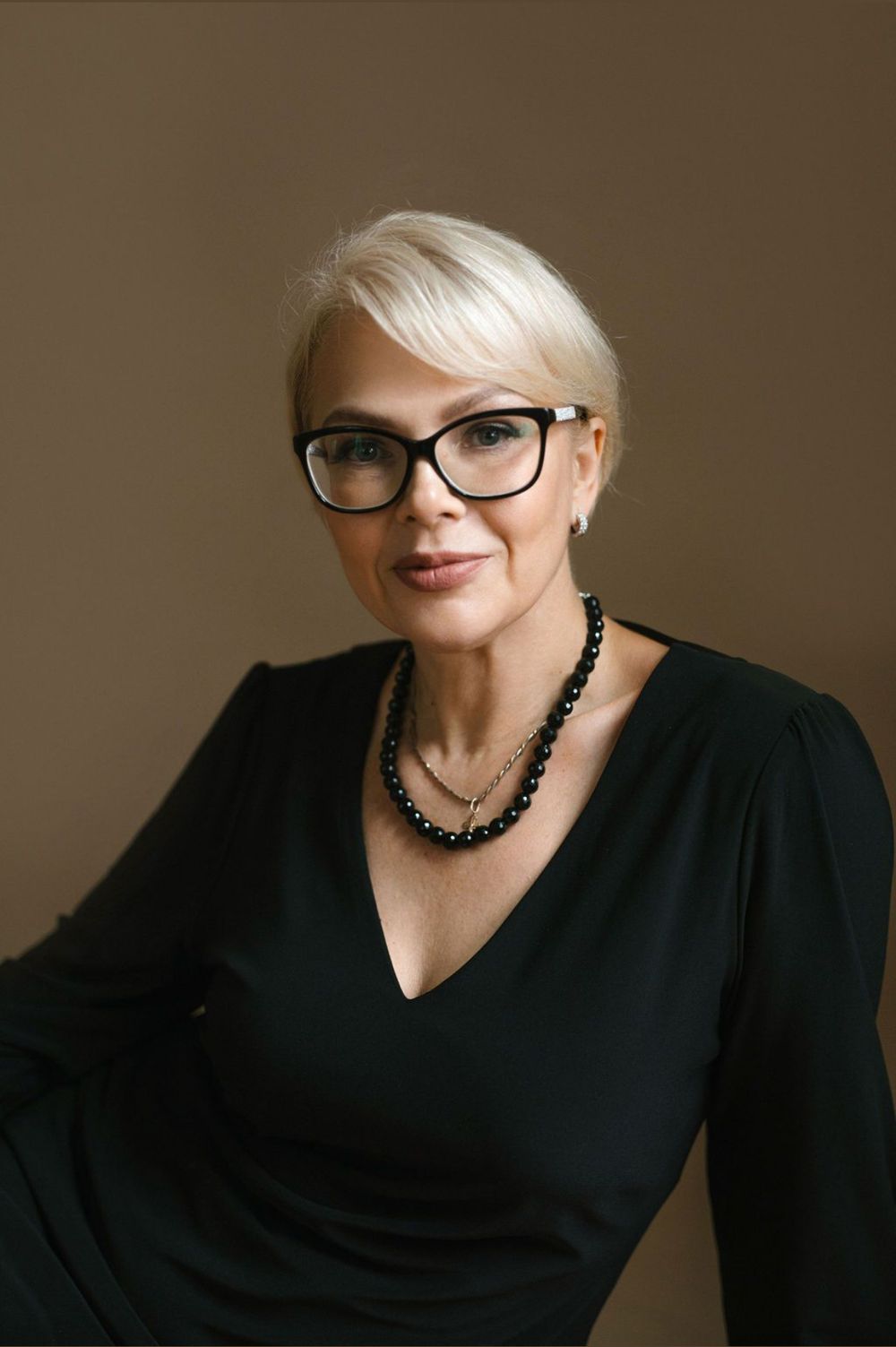Portrait series of someone with platinum blonde hair, black glasses and clothing against a neutral background.