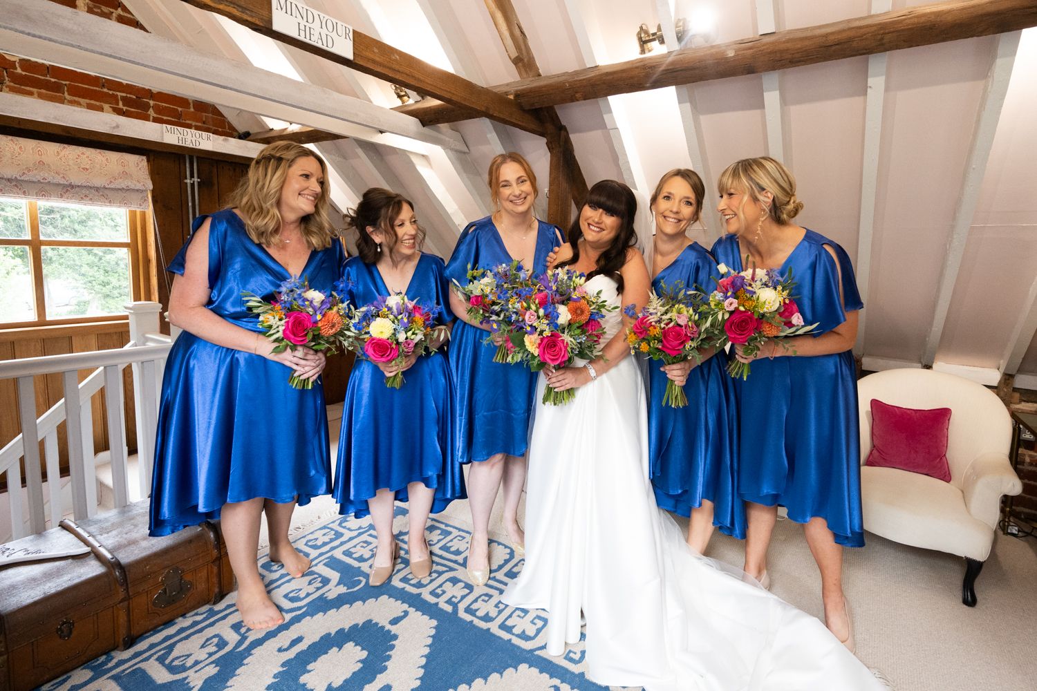 Wedding party in bright blue dresses and white gown gather together in a rustic indoor venue with wooden beams.