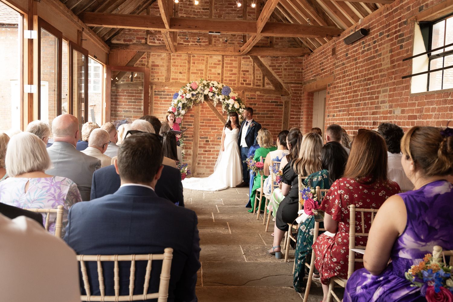 Wedding guests seated in rustic barn venue with exposed wooden beams and brick walls during ceremony.
