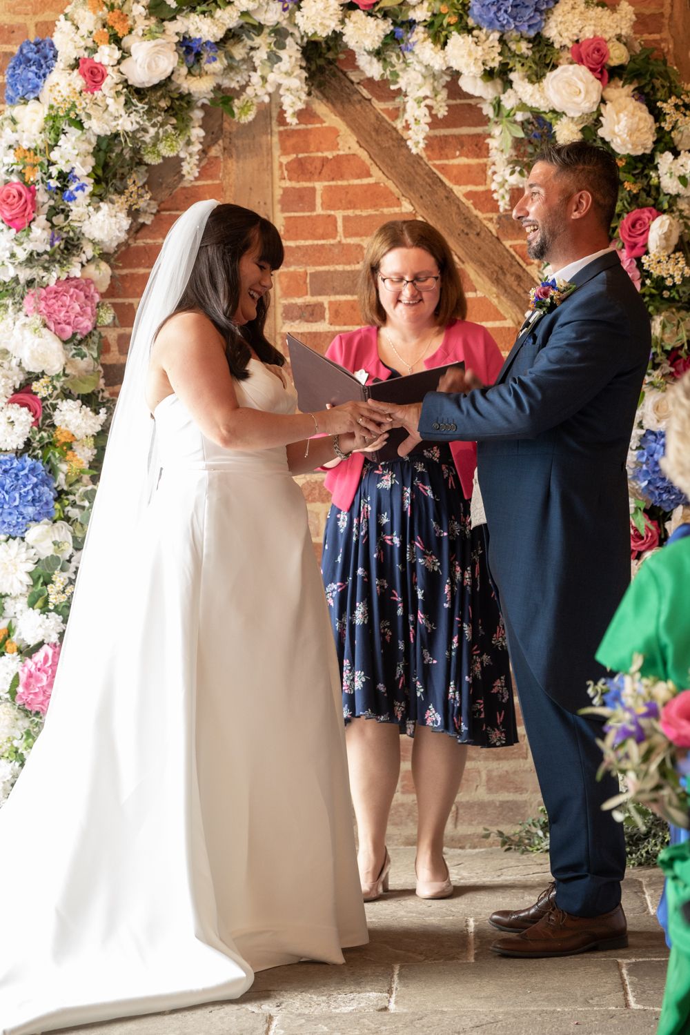 A wedding ceremony takes place under a floral arch with pink, blue and white blooms against a brick wall.