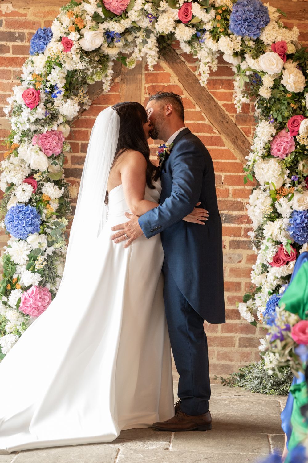 A couple shares their first kiss under a stunning floral arch with pink, white and blue flowers against a brick wall.