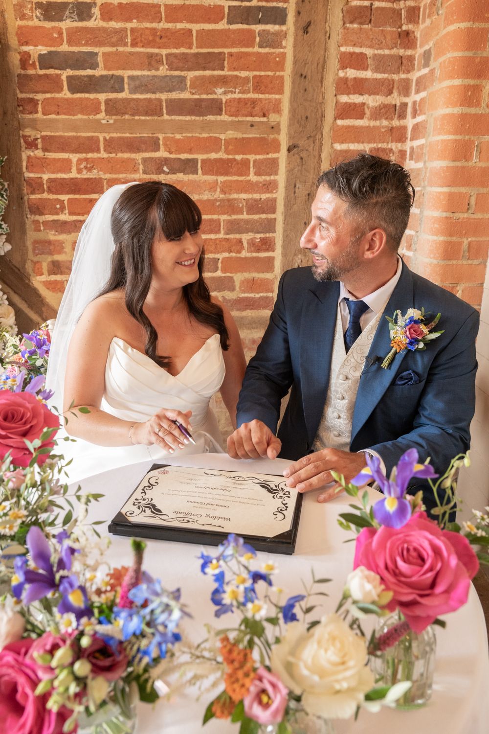 Wedding couple signs marriage certificate at rustic brick venue with colorful floral arrangements on table.