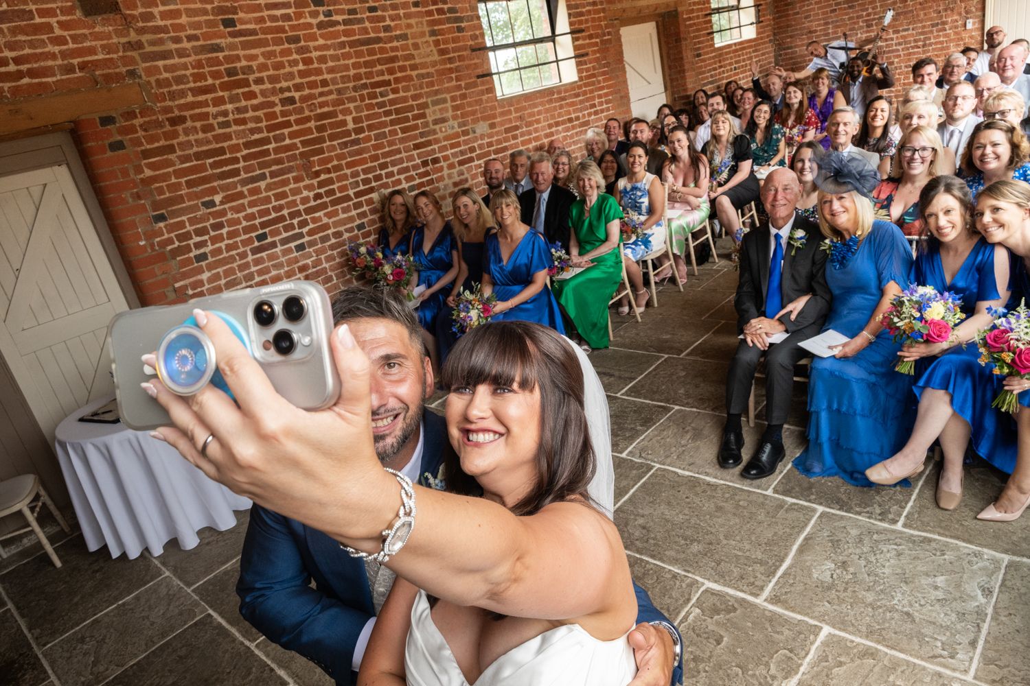 Wedding guests react with joy and excitement during an emotional moment in a rustic brick-walled venue.