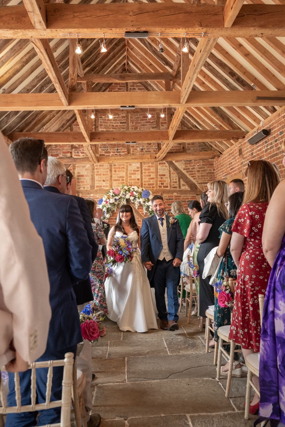 Wedding guests gather inside a rustic barn venue with exposed wooden beams and brick walls during a ceremony.