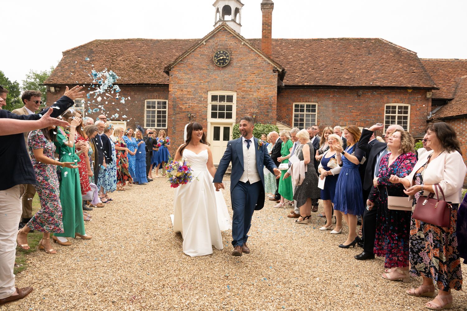 A panoramic view of wedding guests gathered outside a rustic brick barn venue during a countryside celebration.
