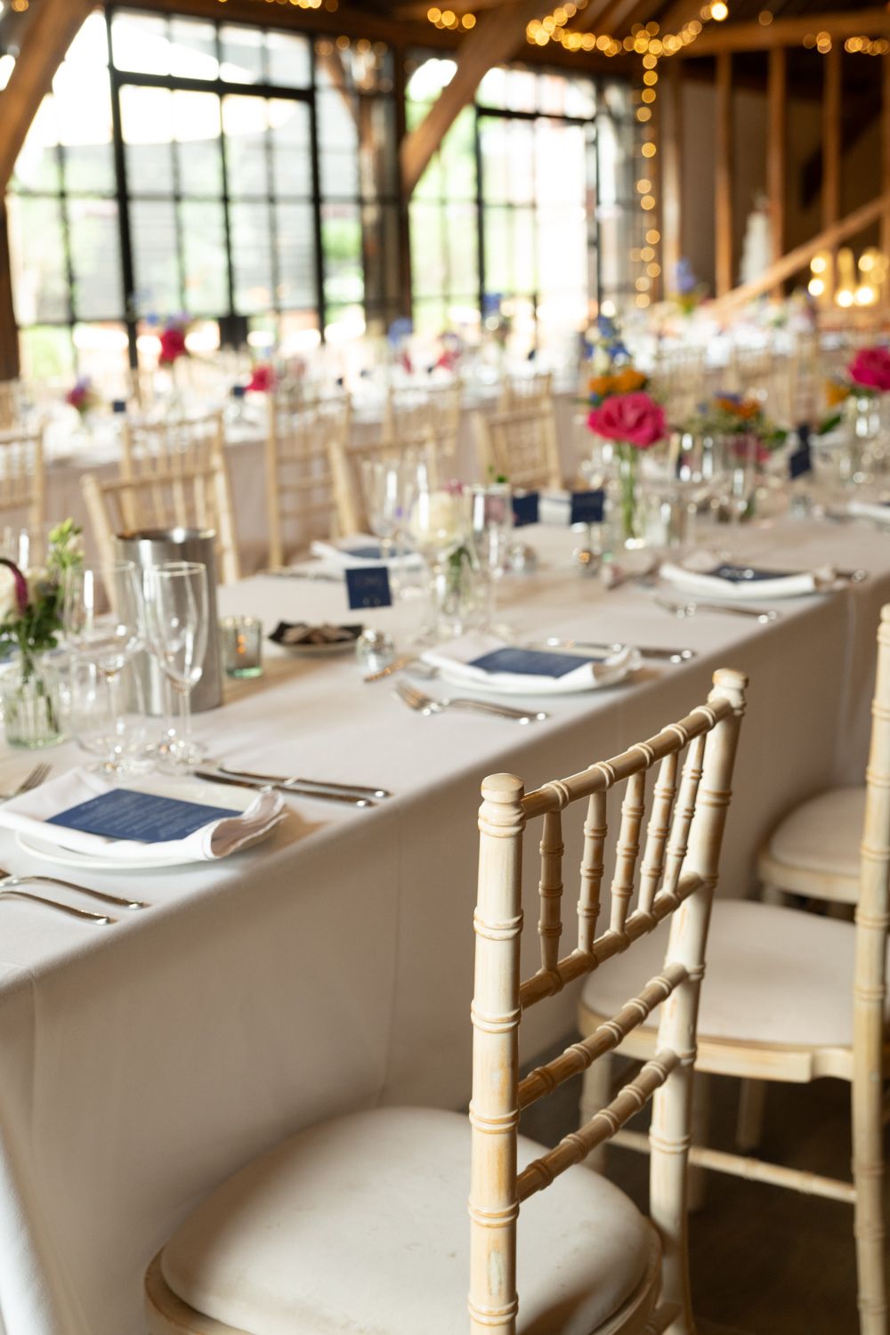 Elegant wedding reception tables set with white linens, gold chiavari chairs, and pink floral centerpieces in a bright venue.