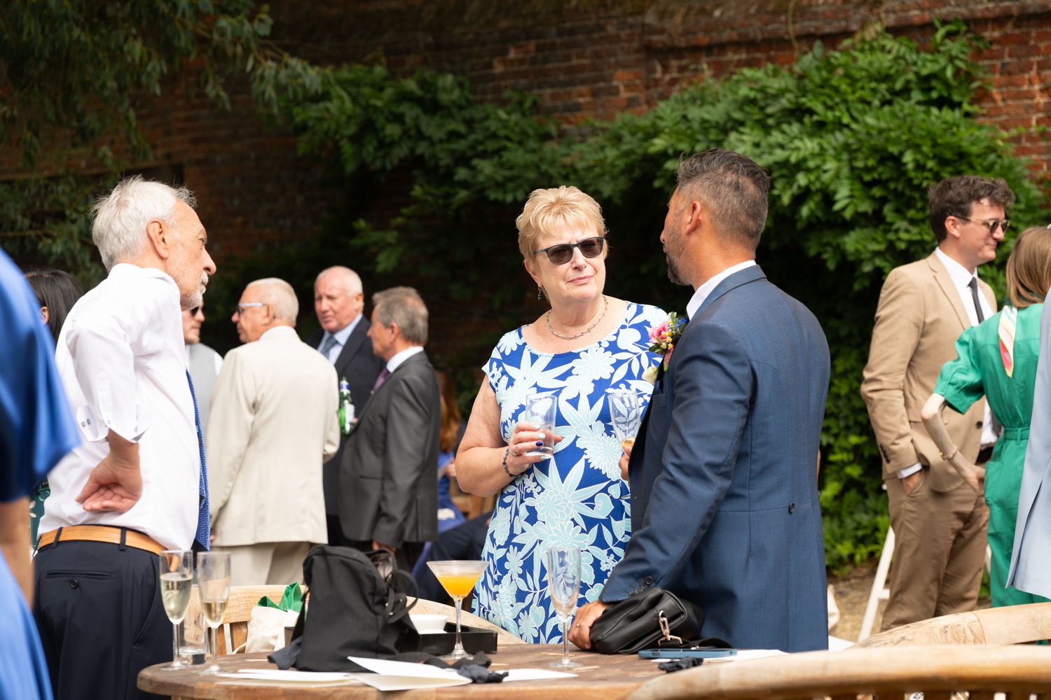Guests mingle and socialize during an outdoor garden party near a brick wall on a sunny day.