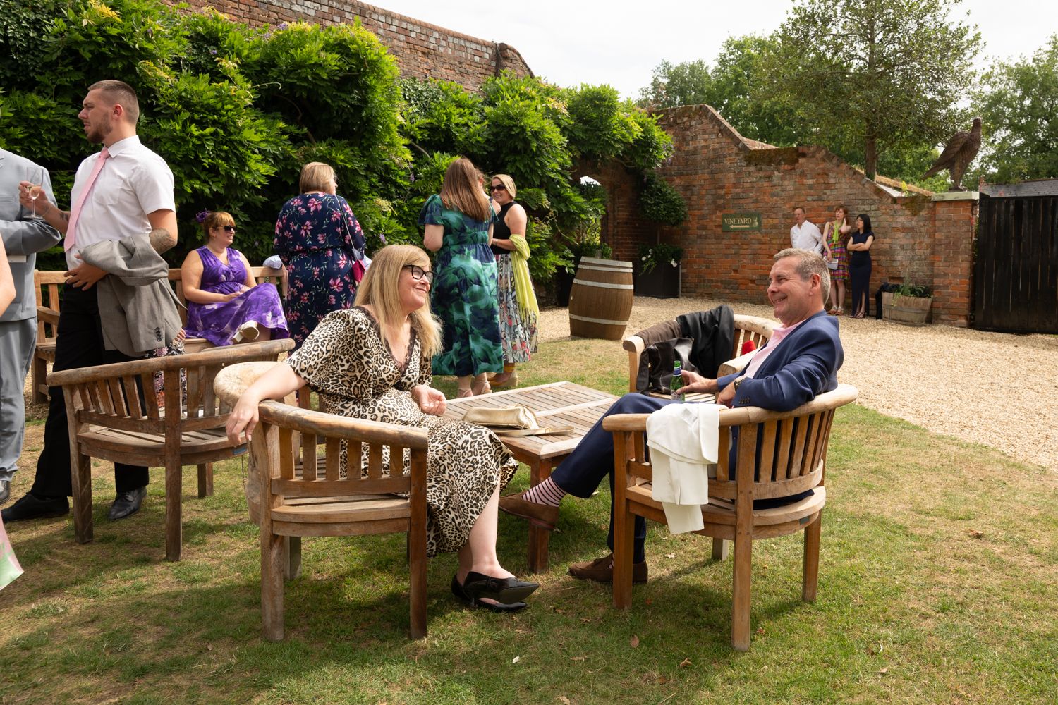 People gather on wooden benches in a garden setting for an outdoor event or gathering.