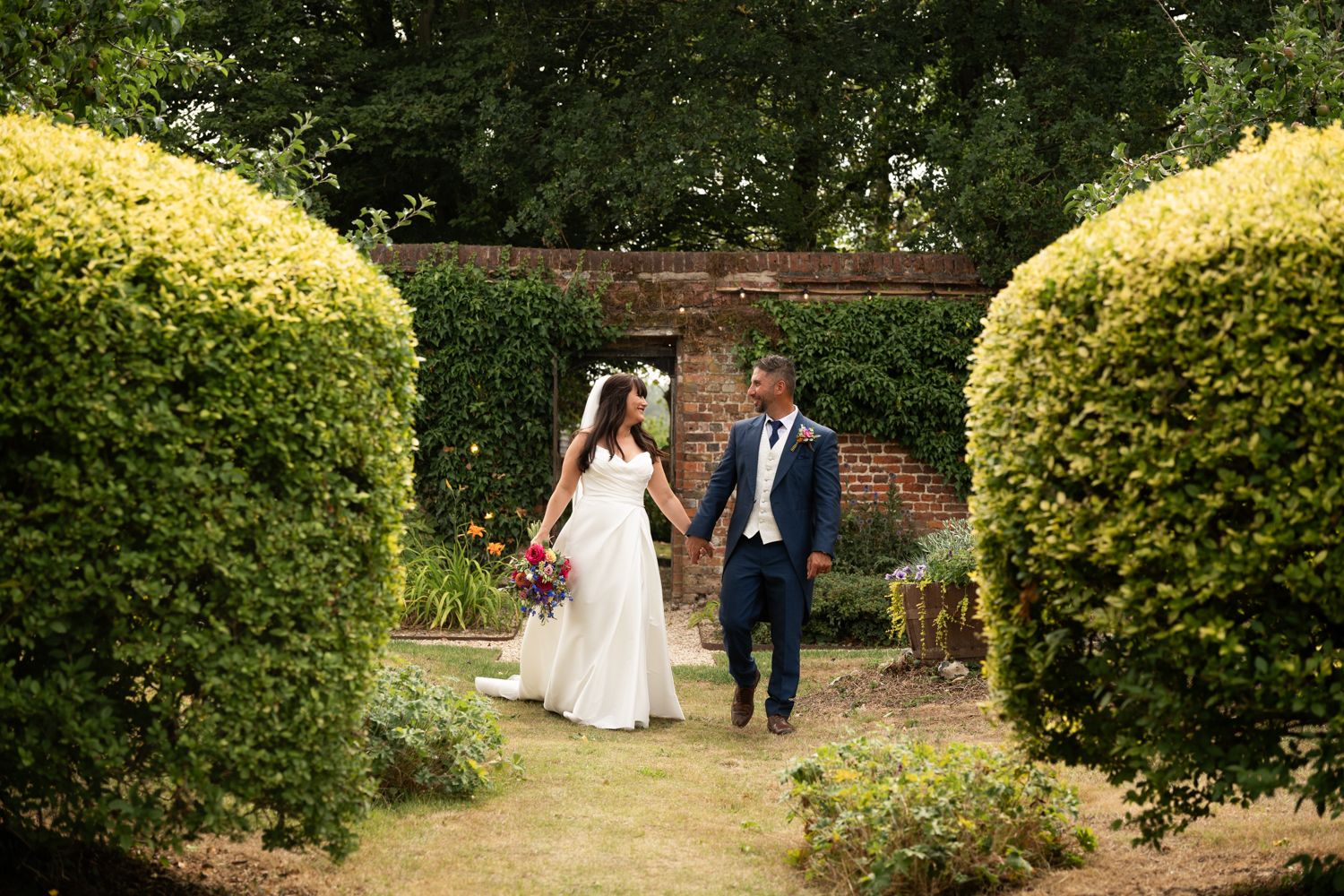 Bride and groom walk hand in hand through a lush garden path with sunlit hedges creating a romantic atmosphere.