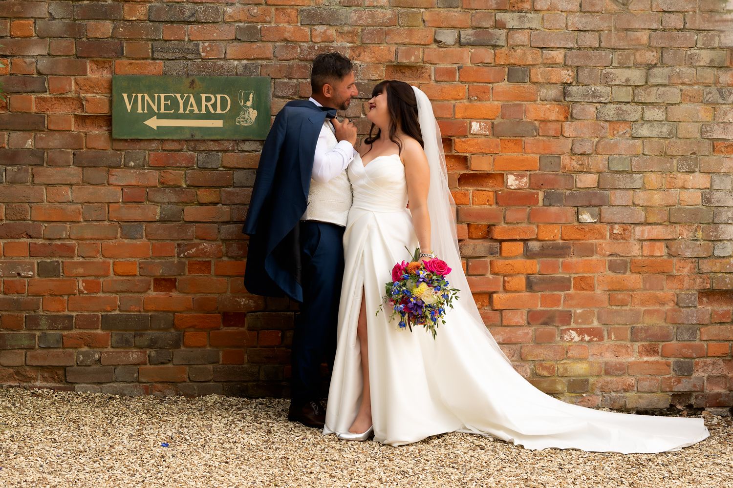 A couple shares a romantic moment against a rustic brick wall with a vineyard sign, the bride holding a vibrant bouquet.