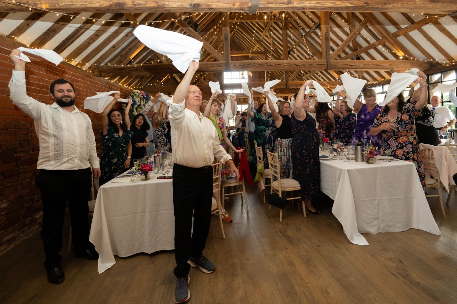 Wedding reception guests celebrate in a rustic barn venue with exposed wooden beams and white decorations.