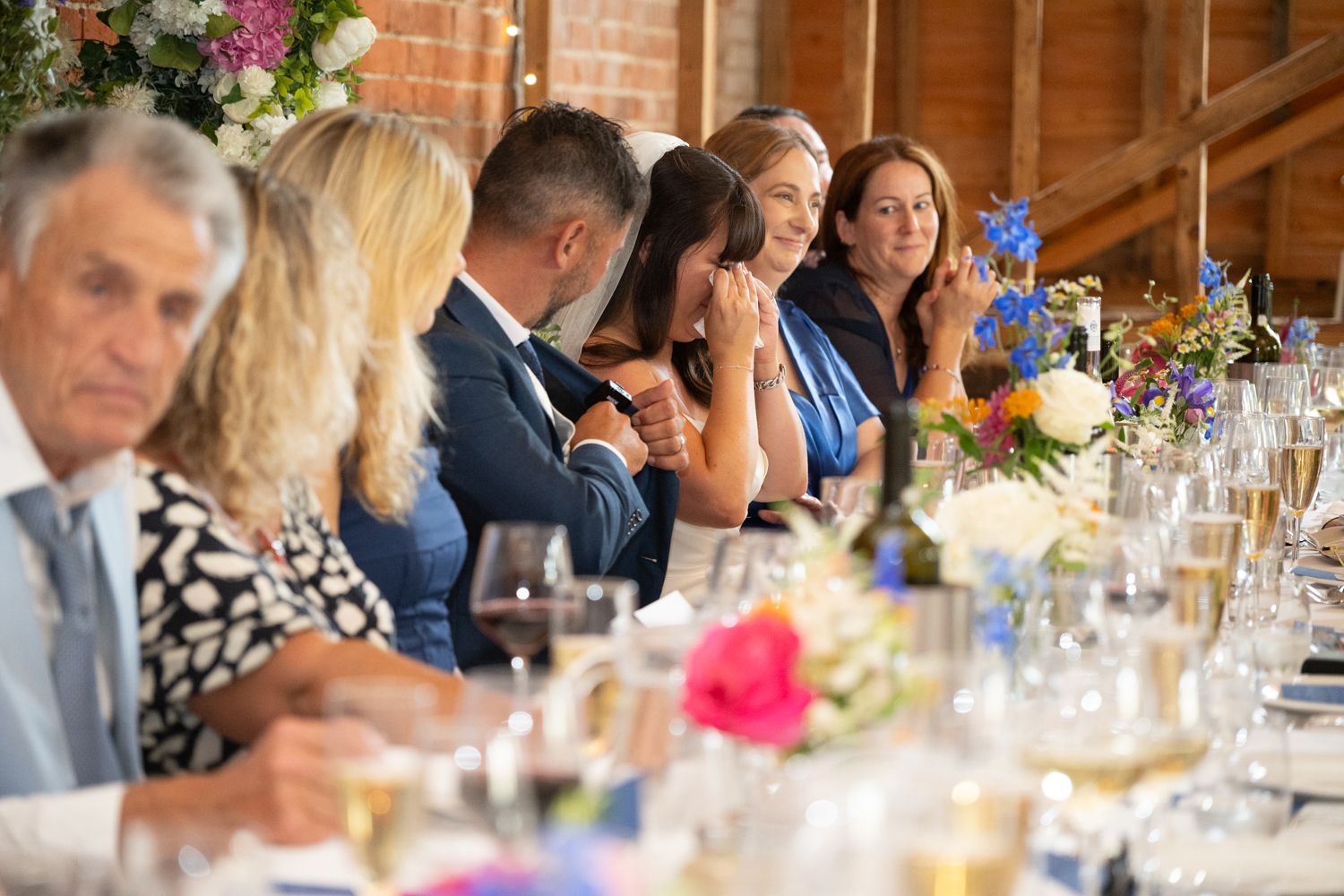 Wedding guests seated at long wooden tables during a rustic barn reception with colorful floral centerpieces.