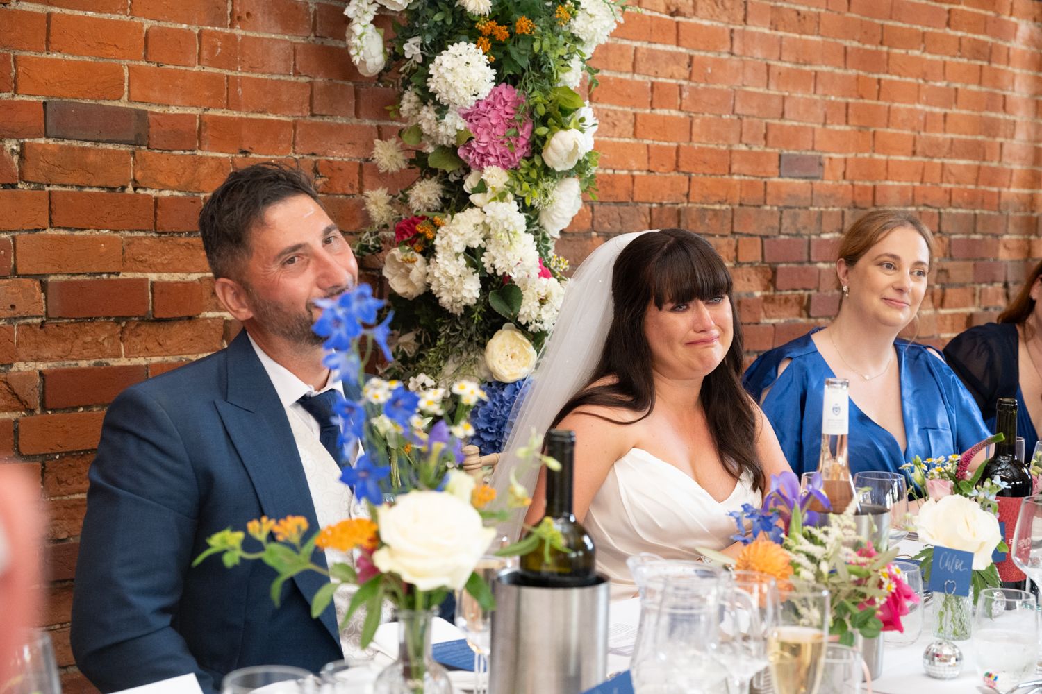 Wedding reception guests seated at long tables with floral centerpieces in a rustic brick-walled venue.