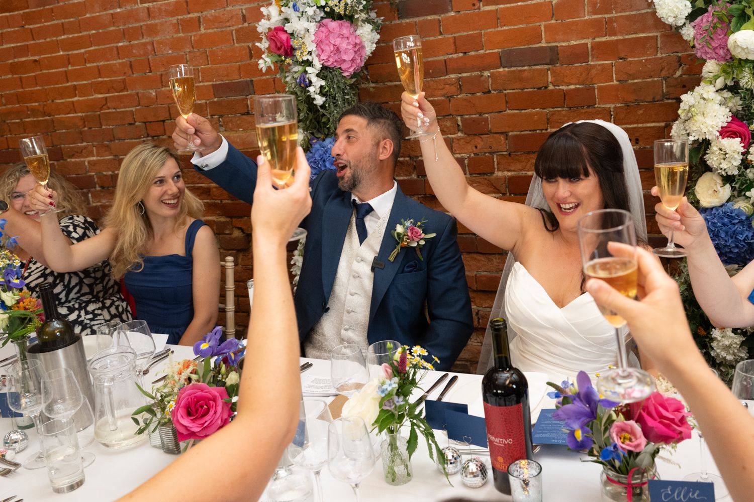 Wedding guests at a rustic brick venue raise their glasses in a celebratory toast during the reception.