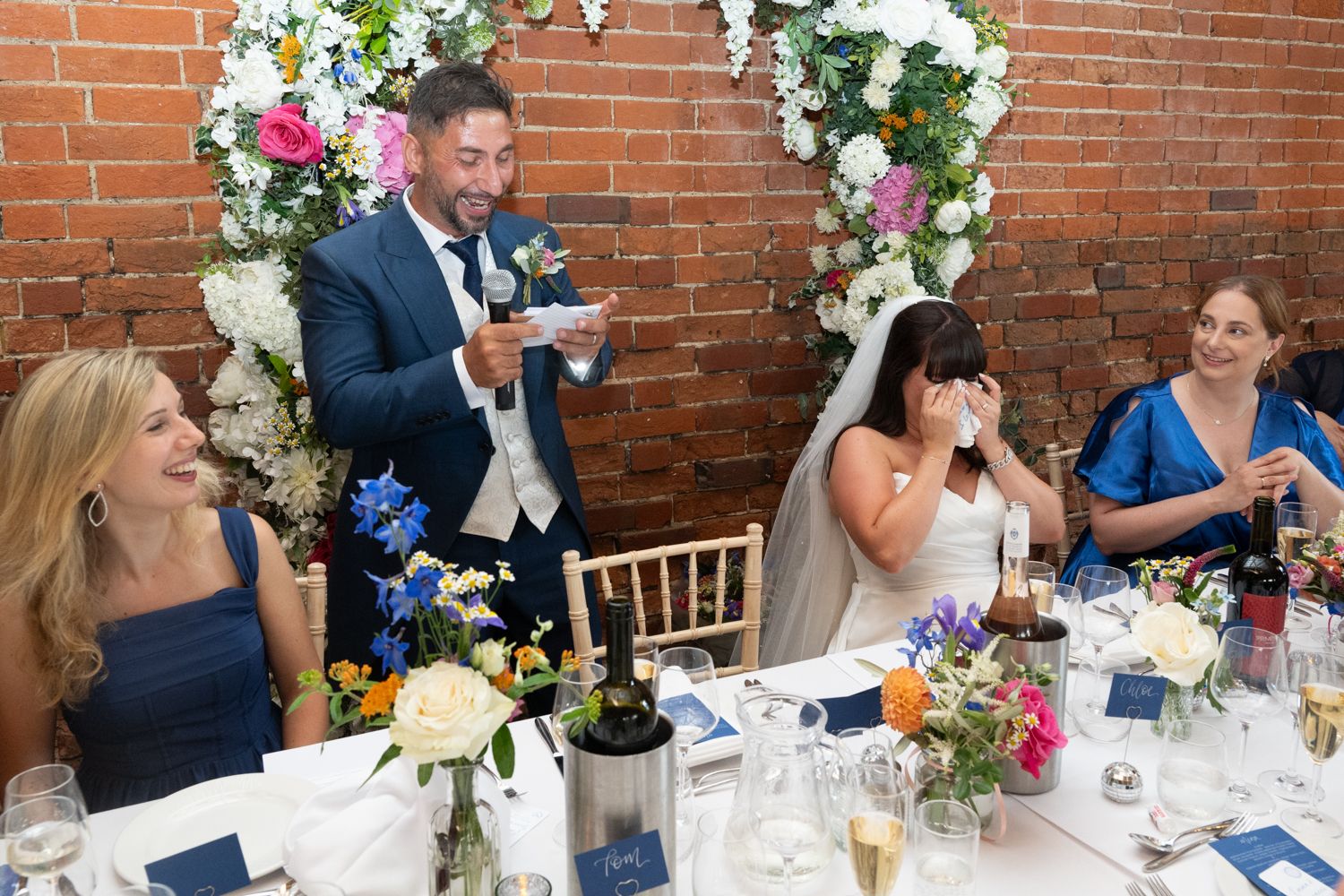 Wedding reception dinner table with guests seated along exposed brick walls decorated with floral arrangements.