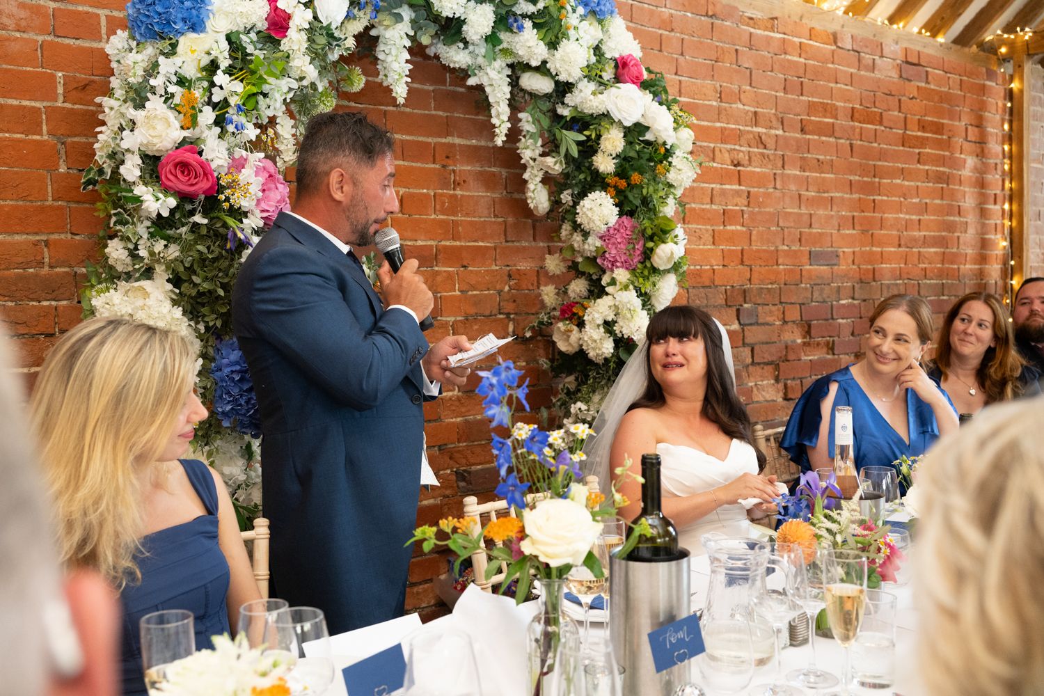 A wedding reception speech being given under a floral arch with white and pink flowers against a brick wall.
