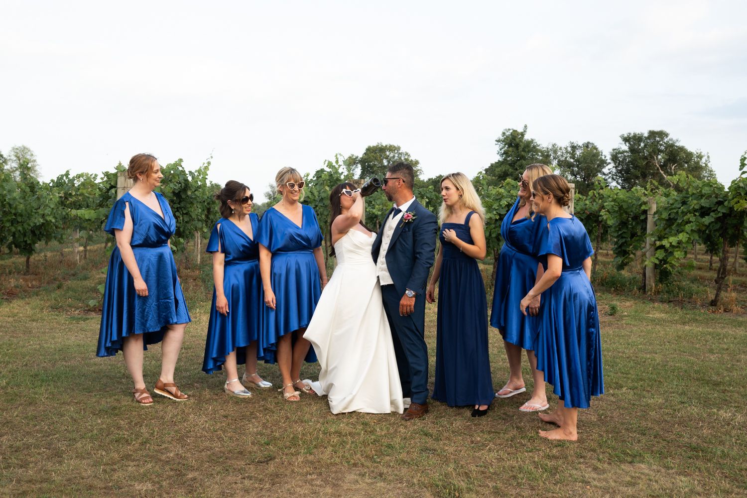 Wedding party in royal blue dresses poses together on a grassy hillside during an outdoor celebration.