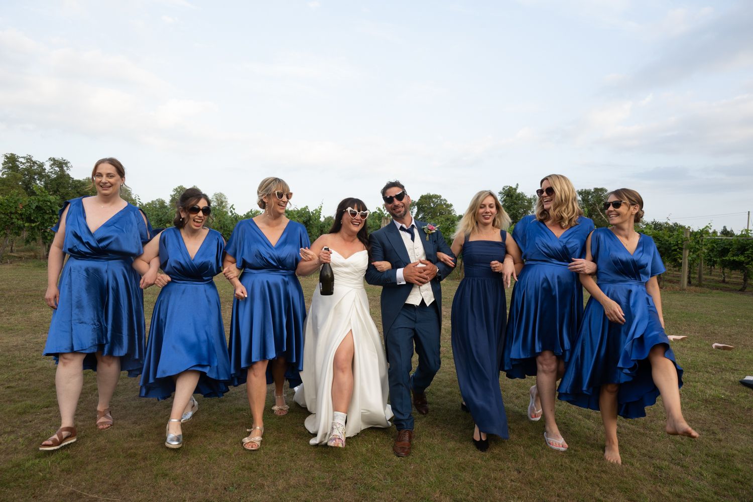 Wedding party in coordinated royal blue dresses poses together on grassy field during outdoor celebration.