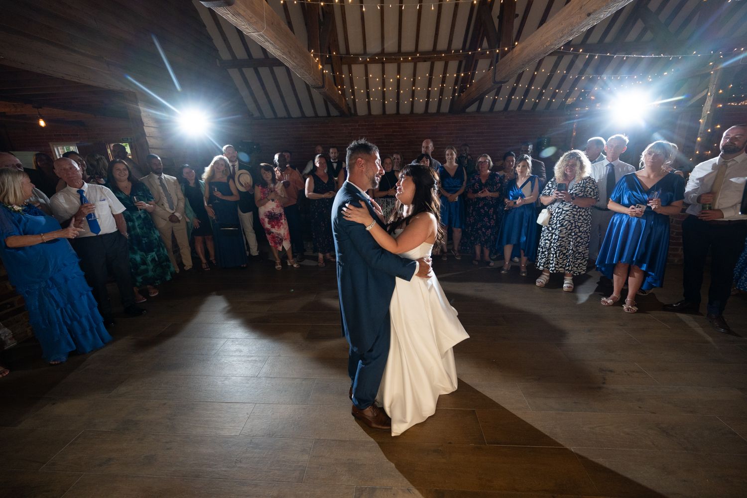 A couple performs their first dance at a rustic wedding reception venue with guests watching from the sidelines.