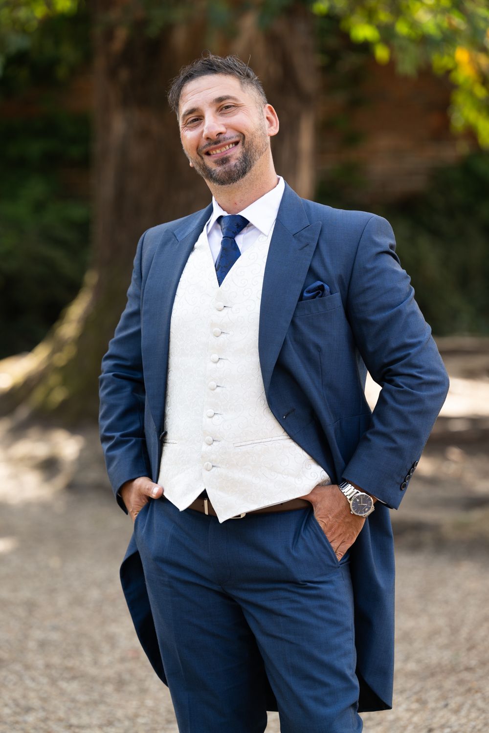 A person in a formal navy blue suit and white vest poses confidently outdoors in a natural setting.