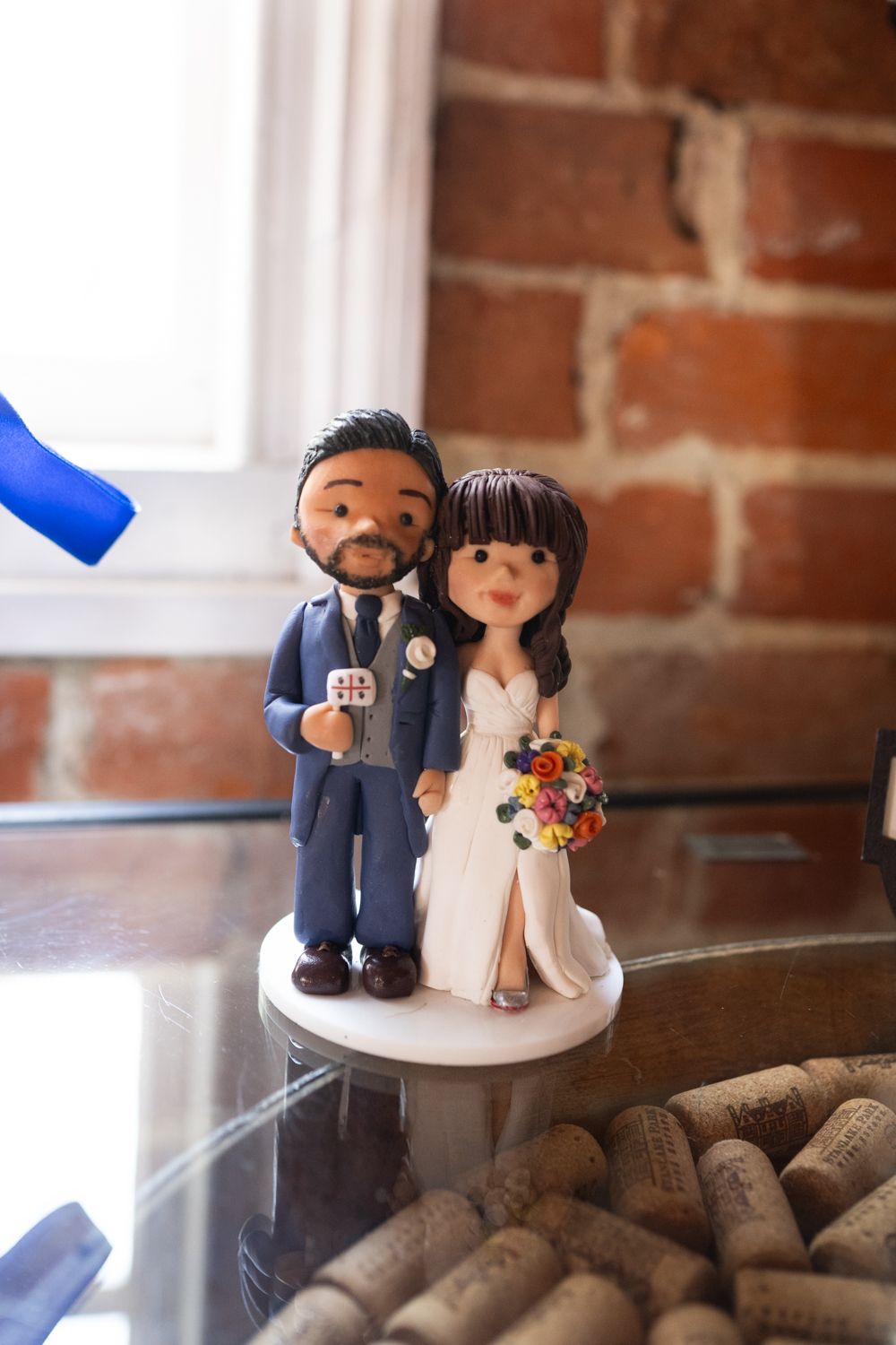 Custom wedding cake topper figurines displayed on a cork surface against a rustic brick wall background.