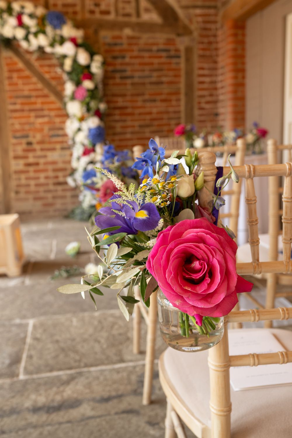 Elegant wedding ceremony aisle decor with pink roses and blue flowers in glass vases on wooden chiavari chairs.