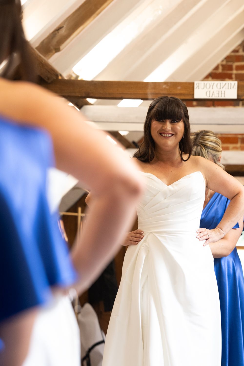 A bride in a white wedding dress gets ready with bridesmaids wearing royal blue dresses in a sunlit room.
