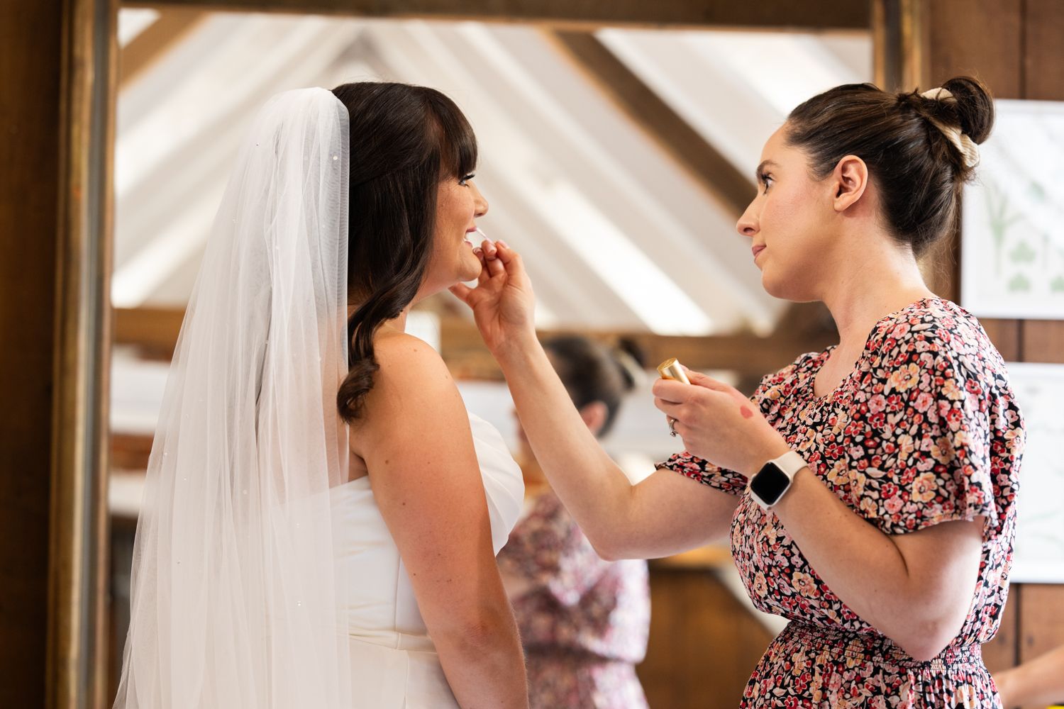 A bride in a white wedding dress gets her makeup touched up by a makeup artist wearing a floral dress.