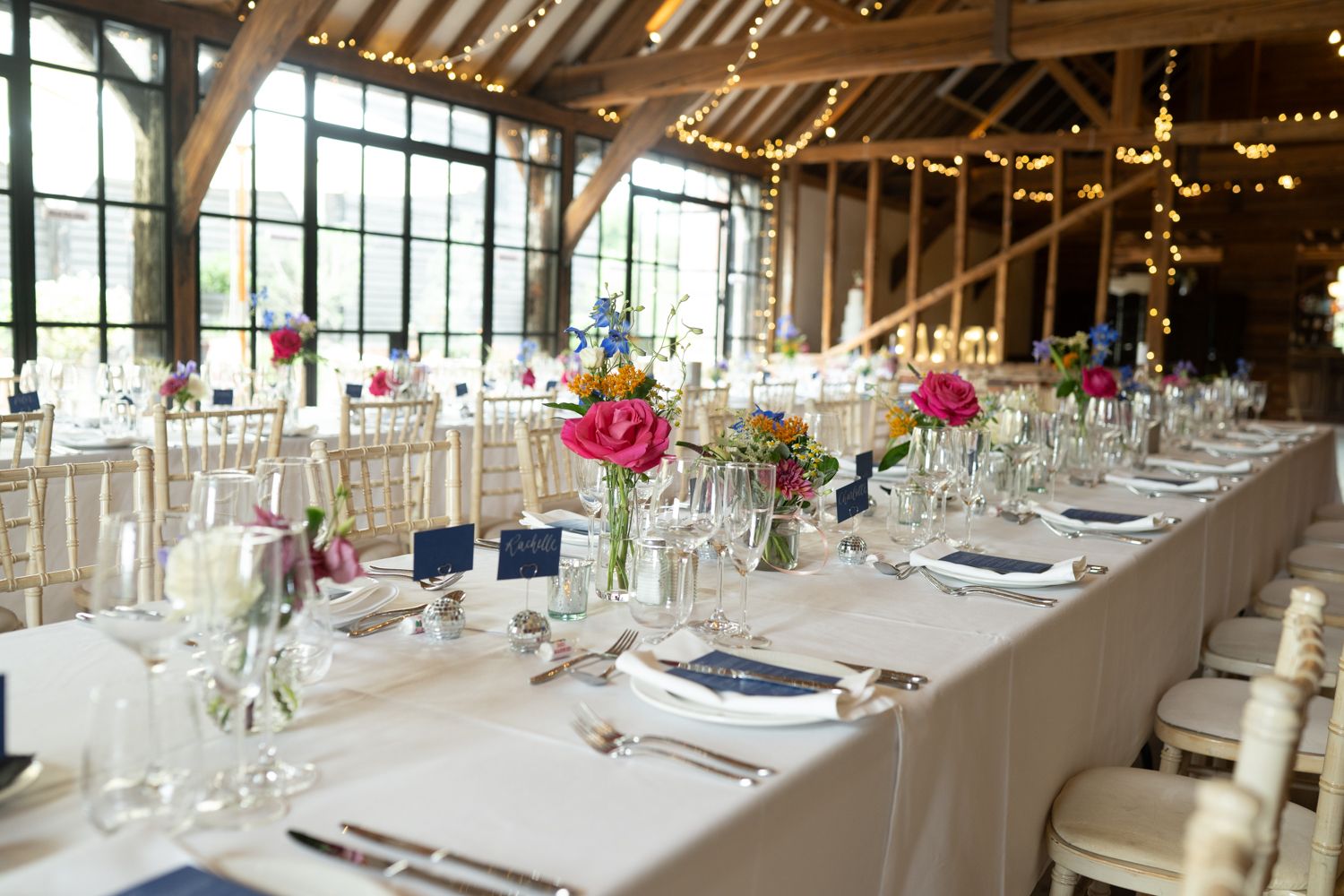 Long banquet tables with white linens and elegant place settings are arranged in a rustic barn venue with exposed beams.