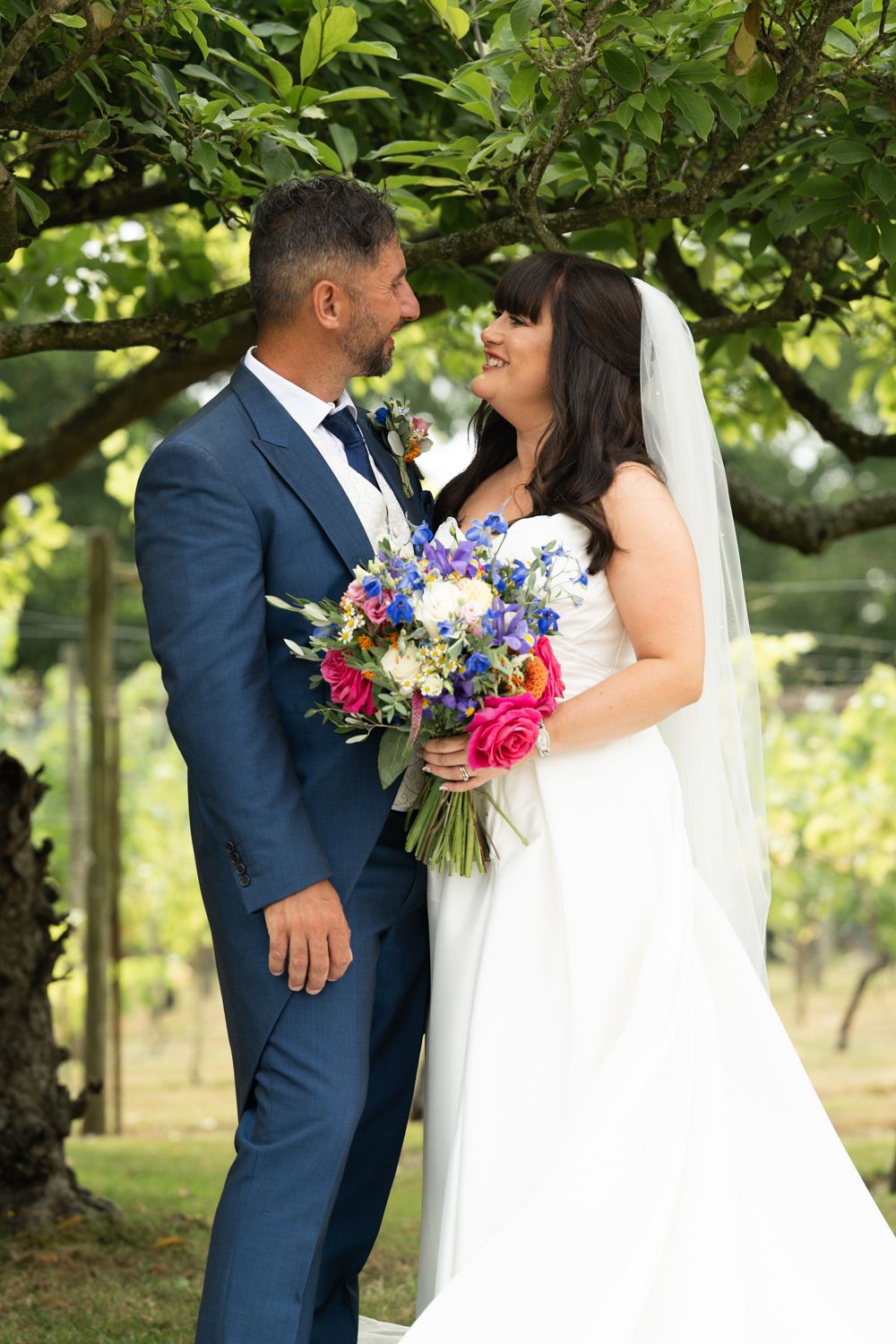 A bride and groom share a romantic moment outdoors under green trees with a vibrant pink and purple bouquet.