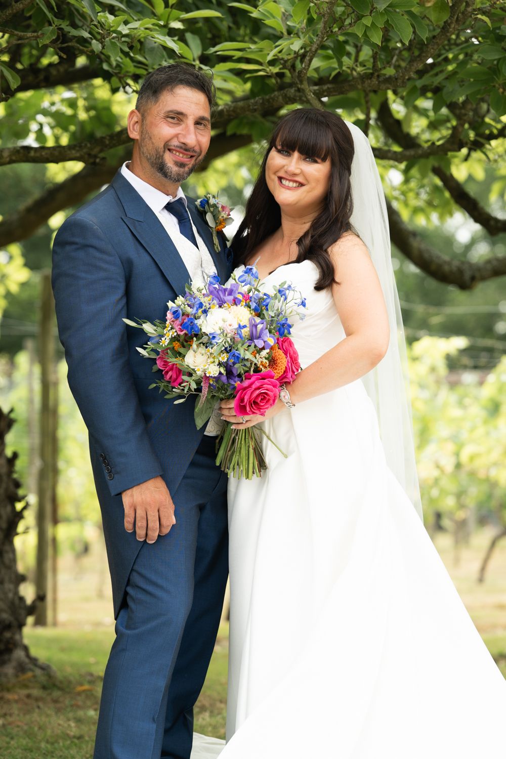 A couple in wedding attire share romantic moments in a lush garden setting with colorful floral bouquet accents.