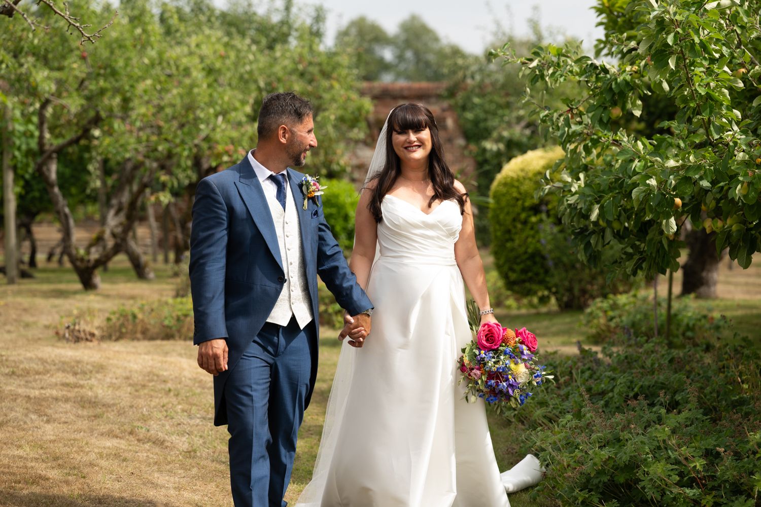 A couple in wedding attire strolls together through a sunlit garden setting on their special day.
