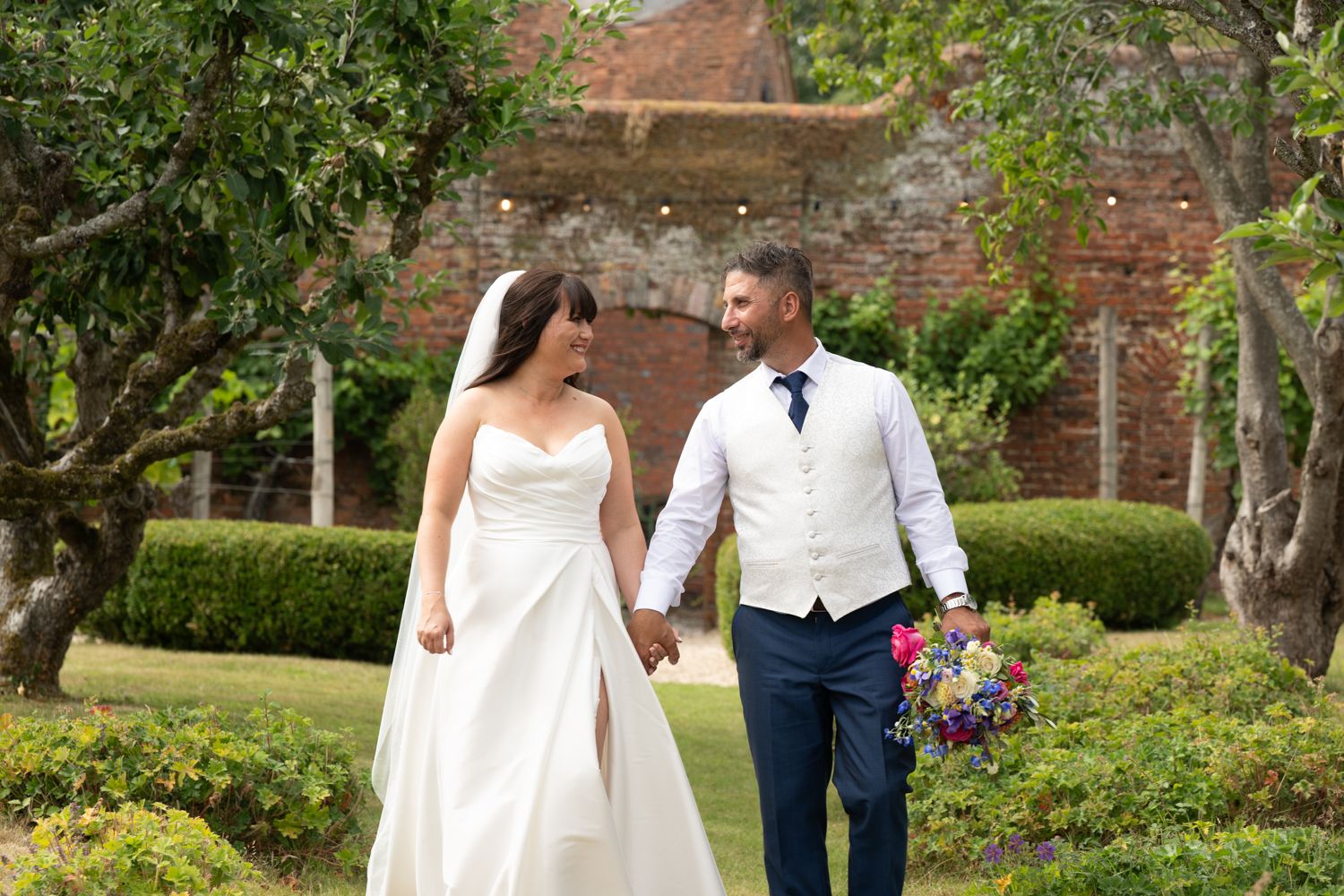 A couple walks hand in hand through a lush garden path with brick walls and greenery surrounding them.