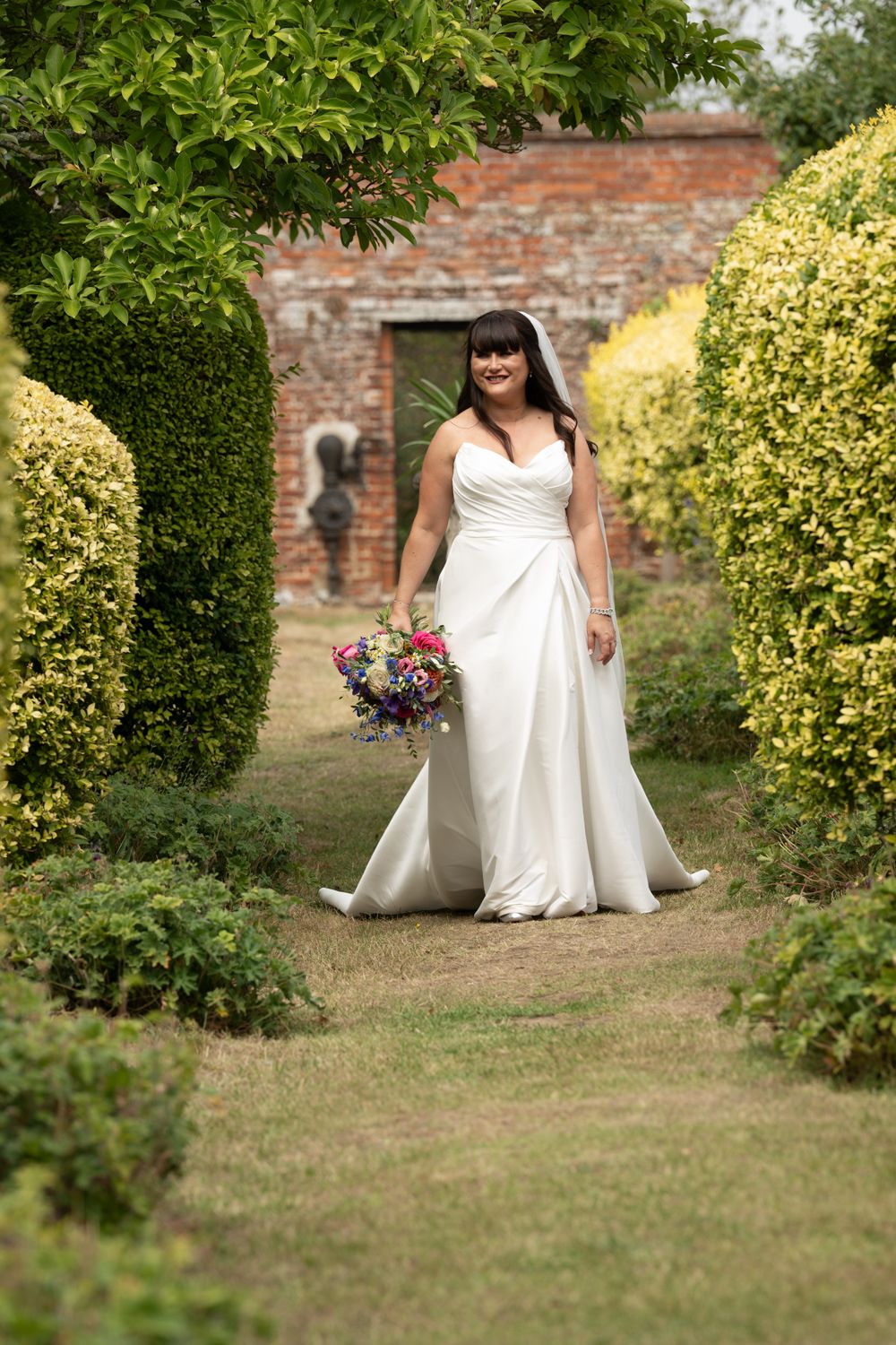 A bride in a white wedding dress poses in a garden setting with yellow flowering bushes and brick walls.