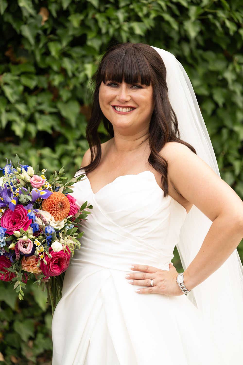 A bride in a white dress holds a vibrant bouquet of pink, blue and purple flowers against a leafy backdrop.