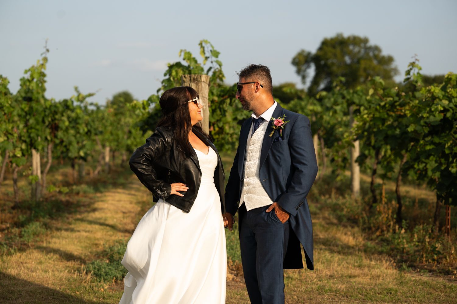 A couple in wedding attire walks through a vineyard on a sunny day with rows of grapevines stretching into the distance.
