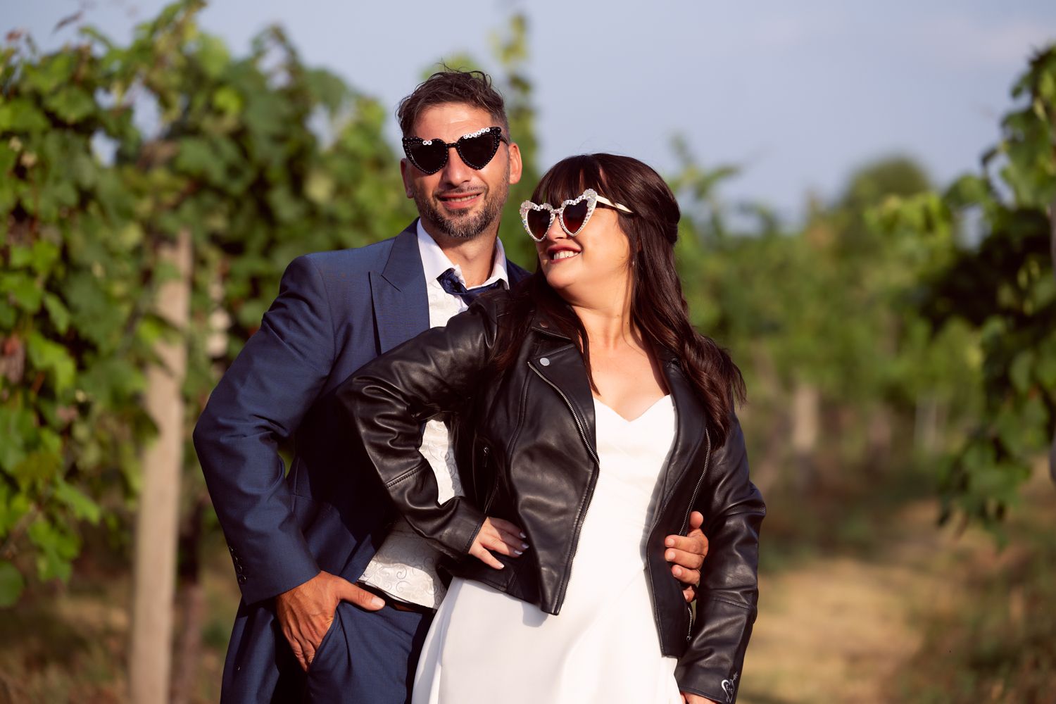 Stylish couple in sunglasses poses together in a sunny vineyard with lush green grapevines in the background.