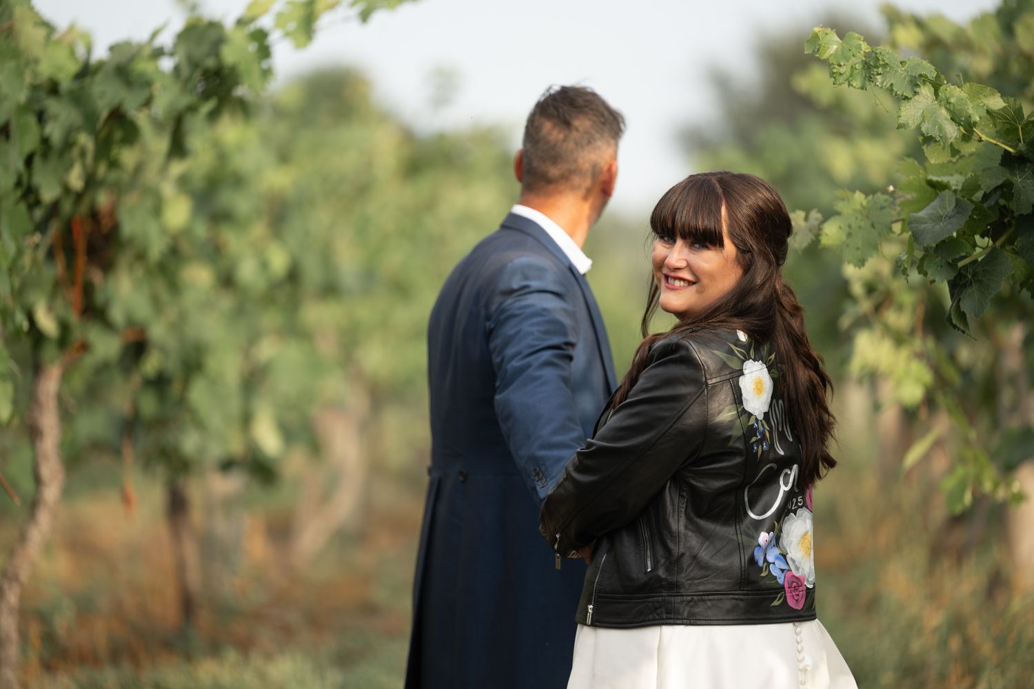 A couple in formal attire stands together in a lush vineyard during a romantic outdoor photoshoot.