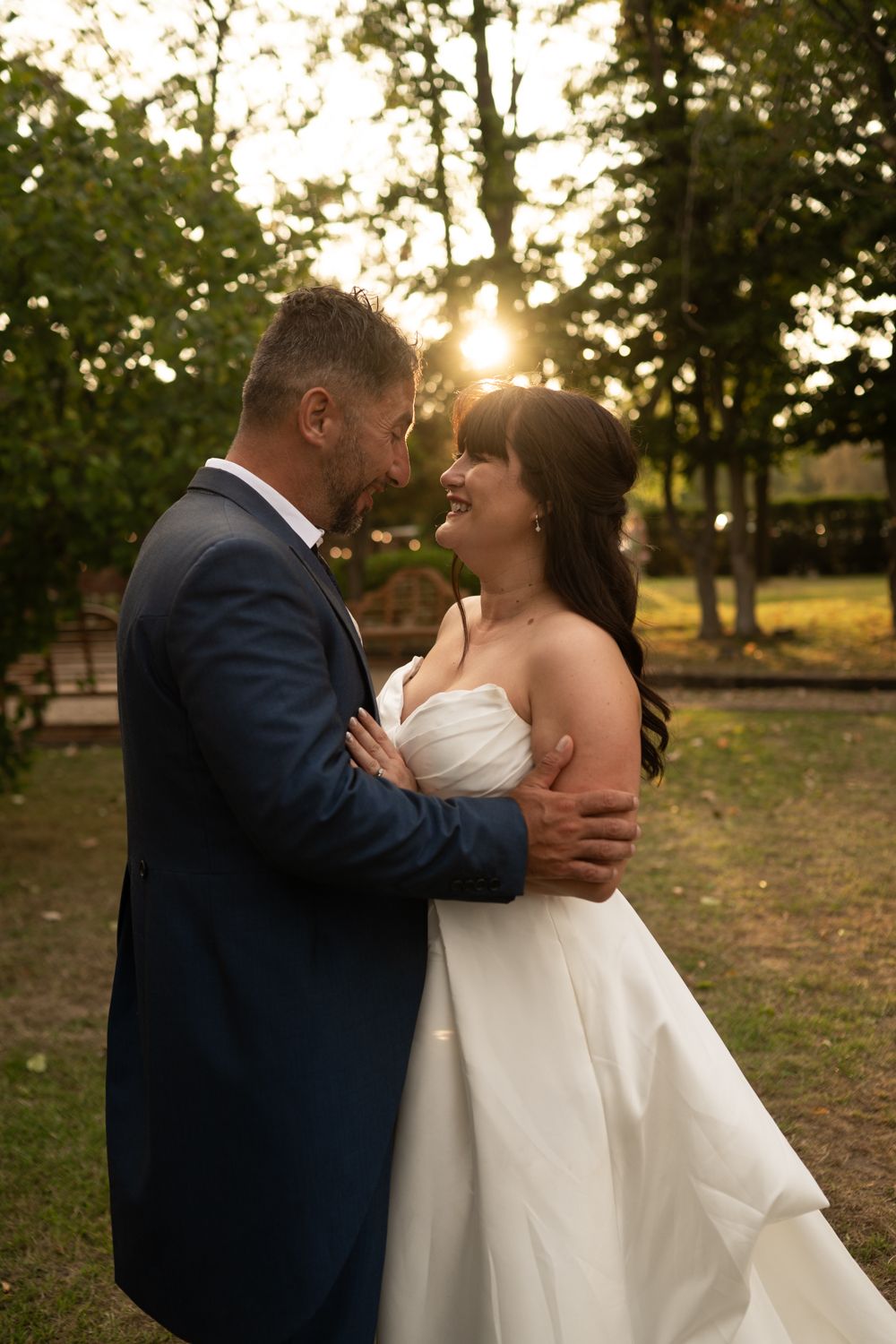 A romantic wedding couple embraces during golden hour in a park setting with sunlight filtering through trees.