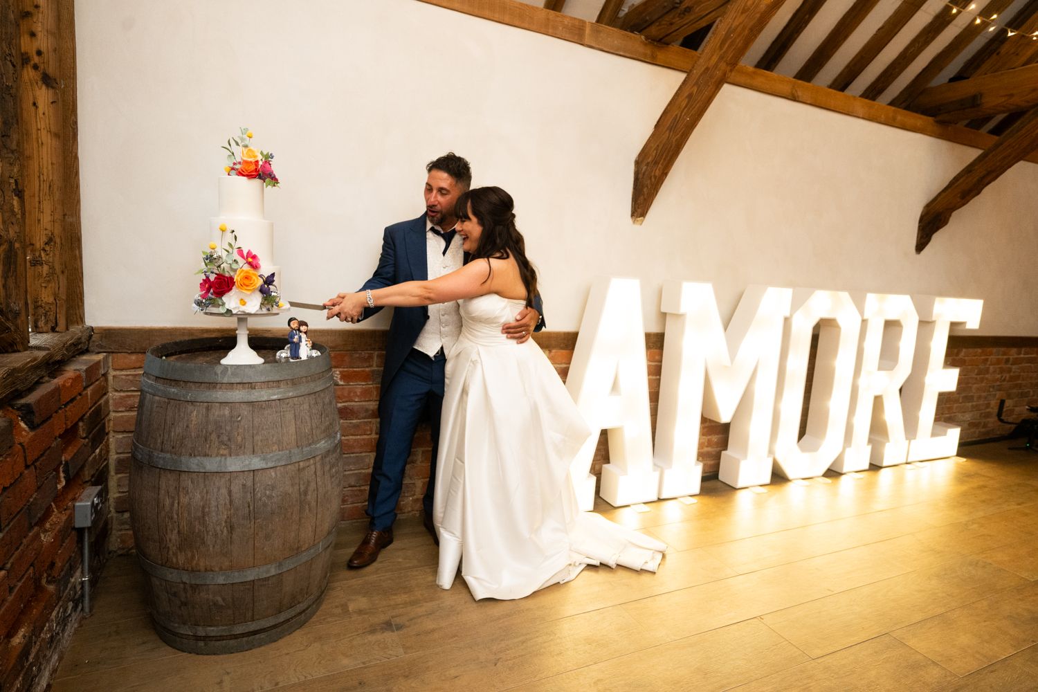 A couple shares a romantic moment in a rustic barn venue with wooden barrels and illuminated letters in the background.
