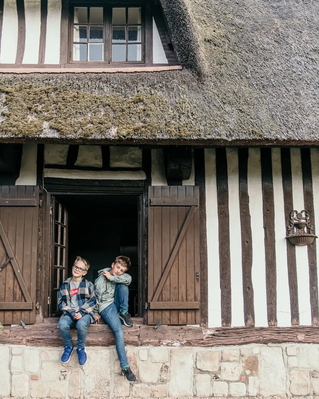 Family sitting on the steps of a timber-frame cottage with a thatched roof in rural France.
