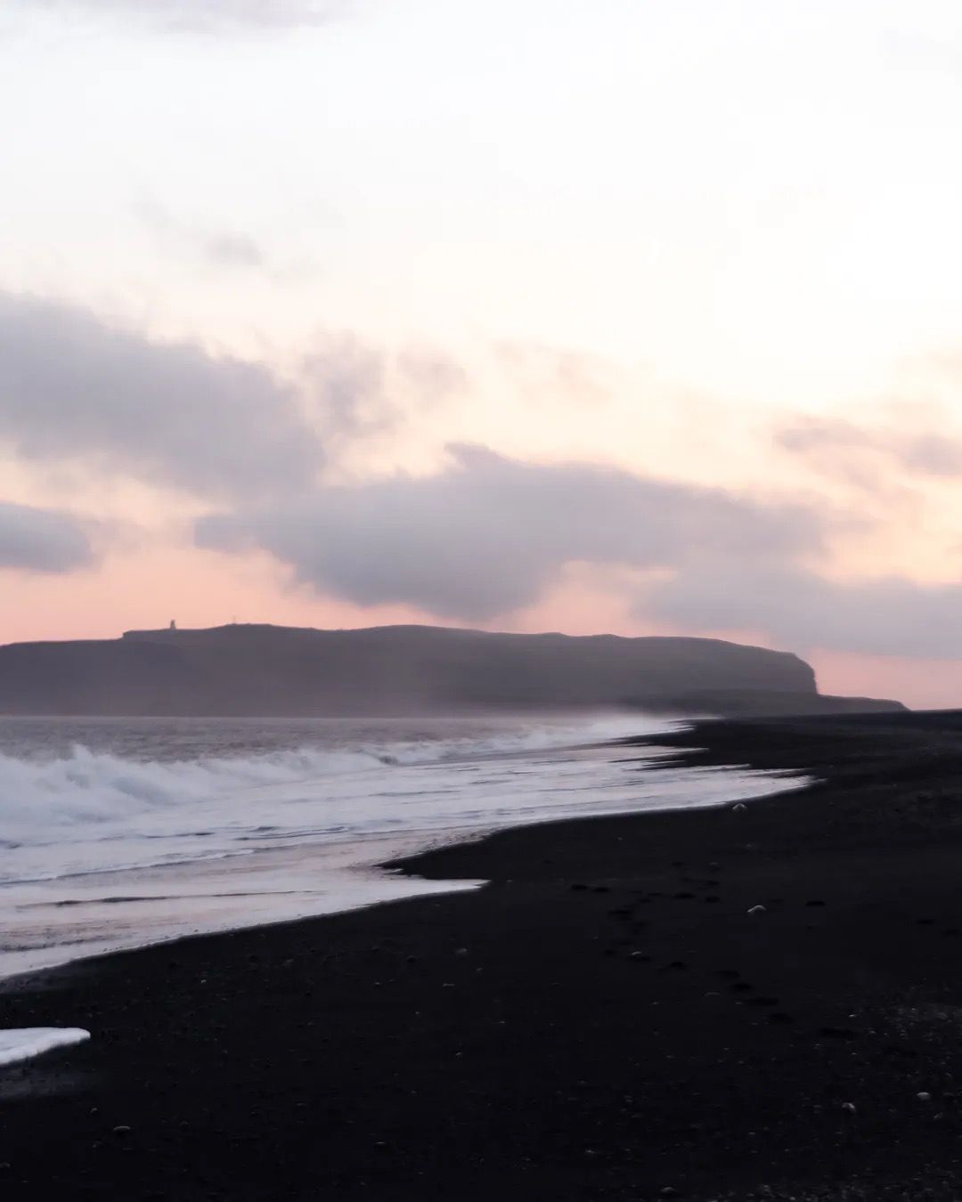 Pink sunset sky reflecting on waves along a black sand shoreline.
