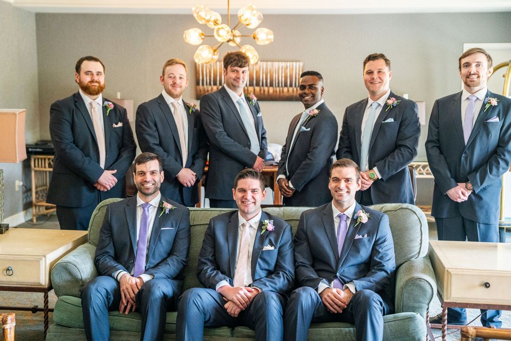 Groomsmen in navy suits sit on gray couches in a hotel lobby while posing for a panoramic wedding photo.
