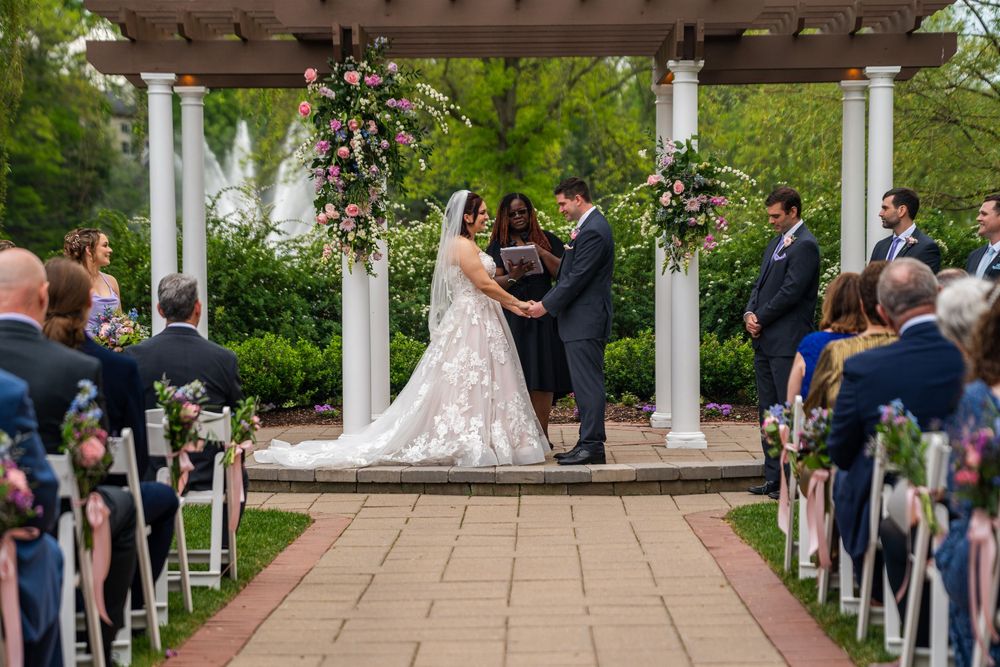 Wedding ceremony takes place outdoors at a garden gazebo venue with guests seated in white chairs.