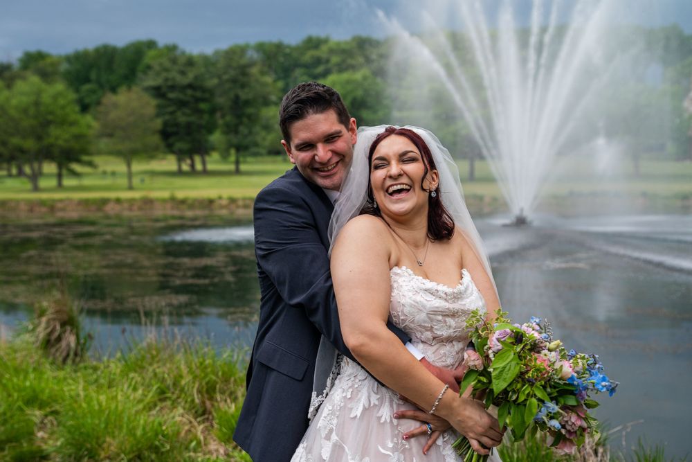 A couple poses for romantic wedding photos near a fountain in a scenic park setting with lush greenery and water features.