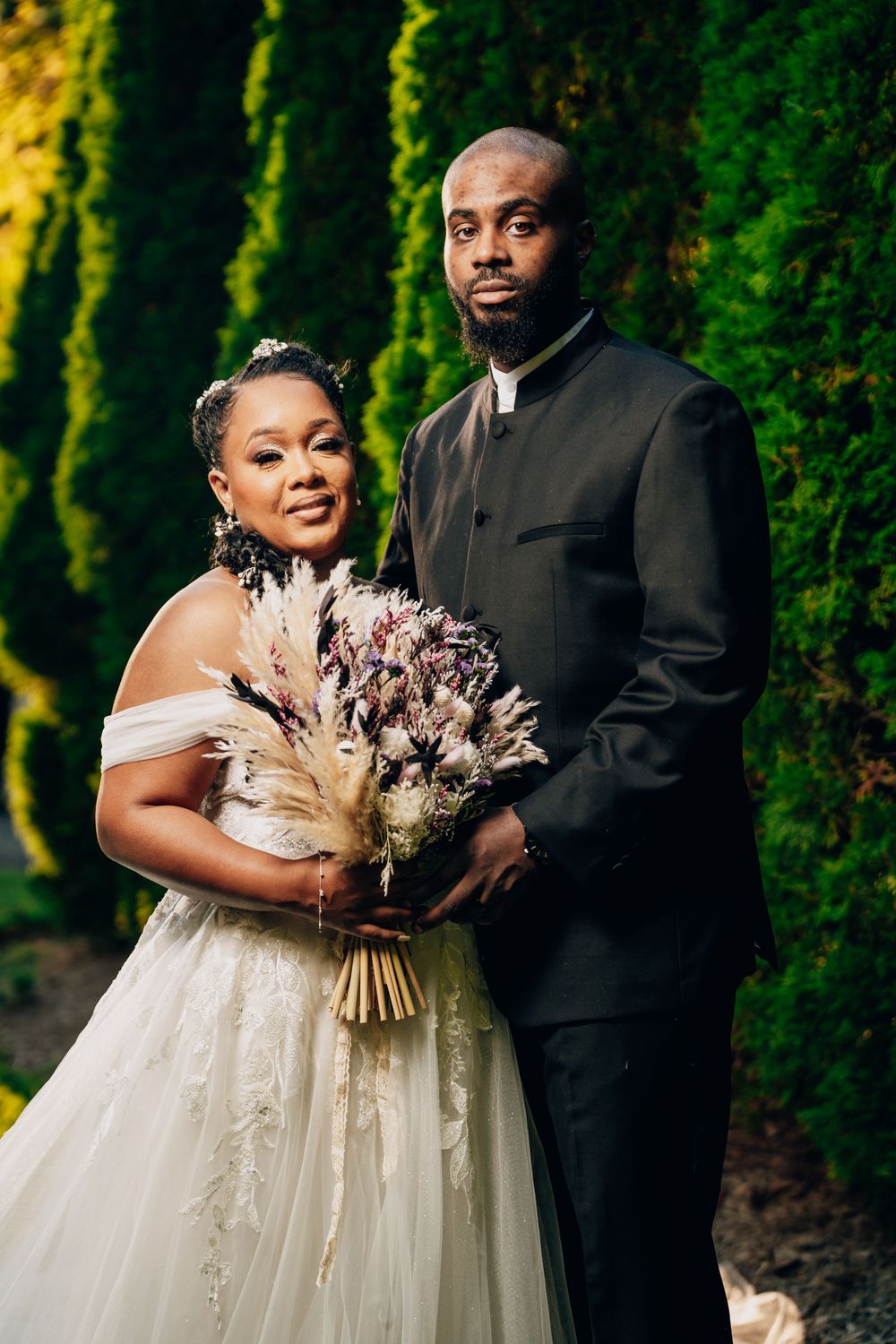 A couple poses for a wedding photo outdoors with a dried flower bouquet against lush green foliage.